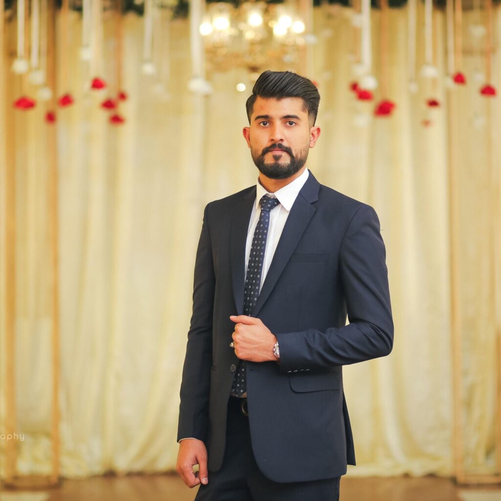 Bearded man in a dark suit poses confidently in a formal indoor setting.